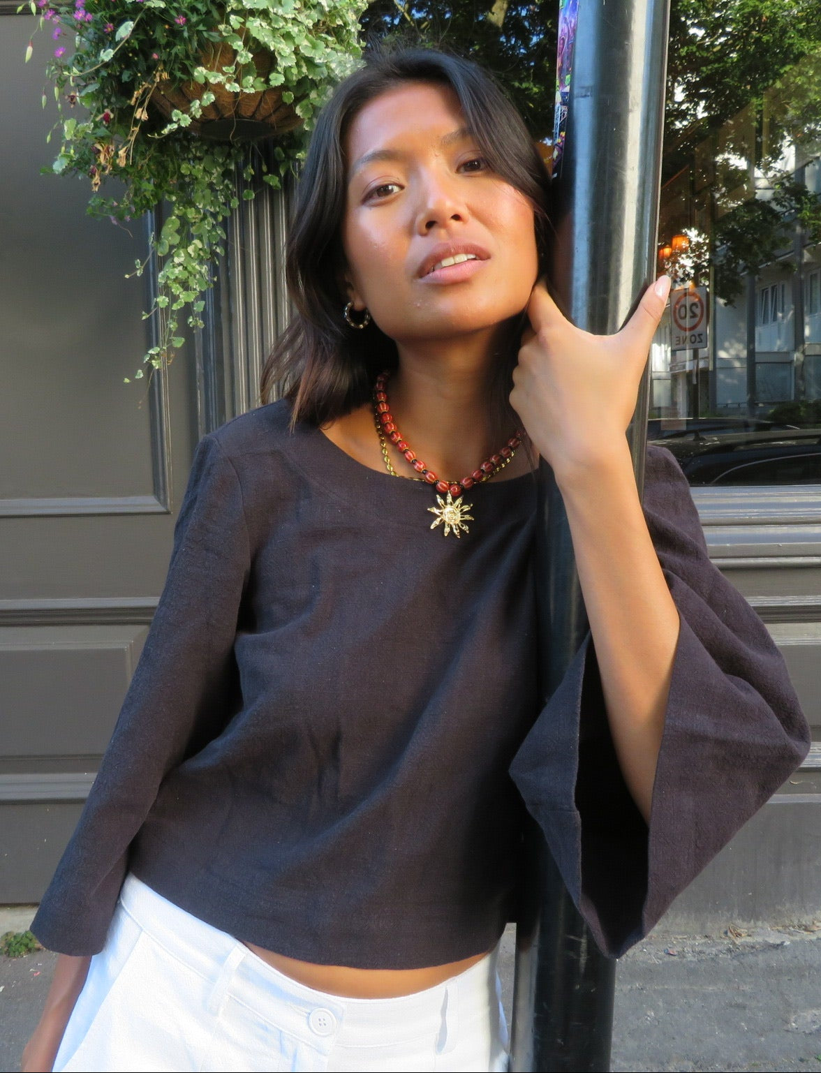 A woman wearing a dark brown cropped top with wide three-quarter sleeves and white pants, posing outdoors next to a black pole in front of a building with a gray door.