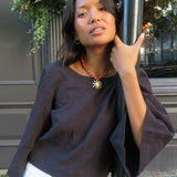 A woman wearing a dark brown cropped top with wide three-quarter sleeves and white pants, posing outdoors next to a black pole in front of a building with a gray door.