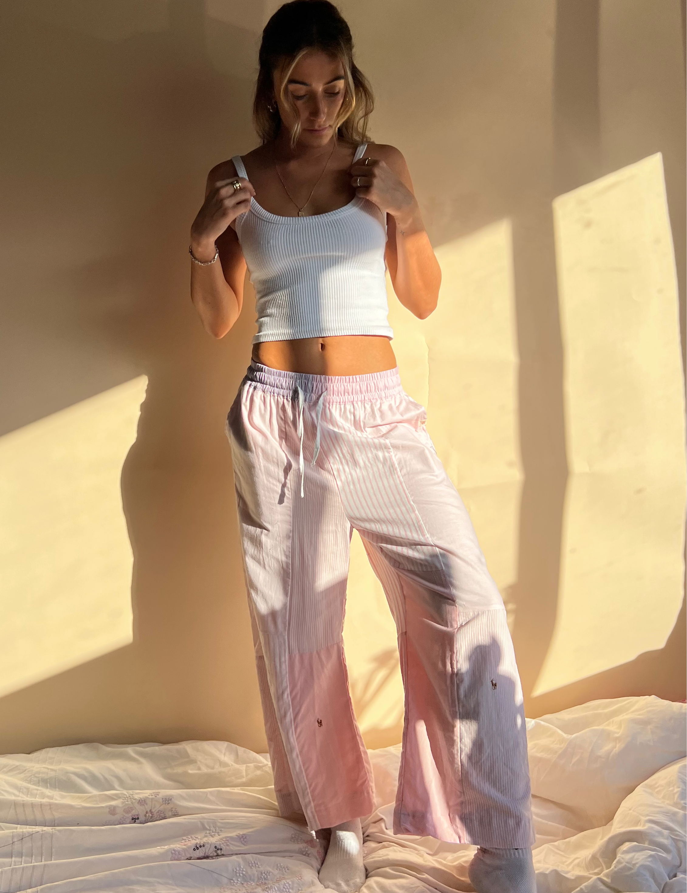 A person wearing baby pink trousers with a wide leg and a white top, standing in a room with sunlight casting a shadow on the wall.