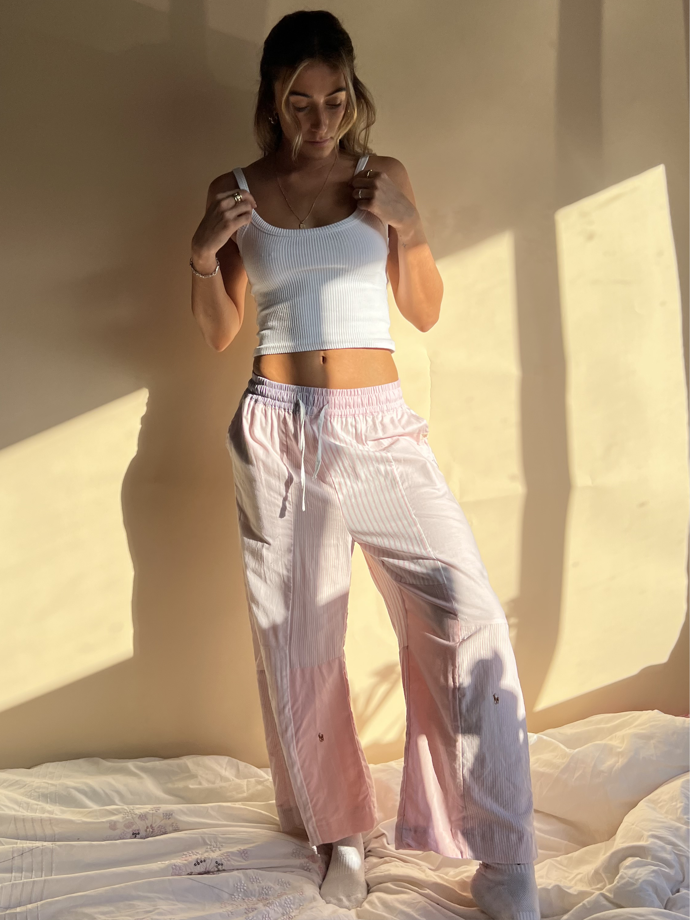 A person wearing baby pink trousers with a wide leg and a white top, standing in a room with sunlight casting a shadow on the wall.