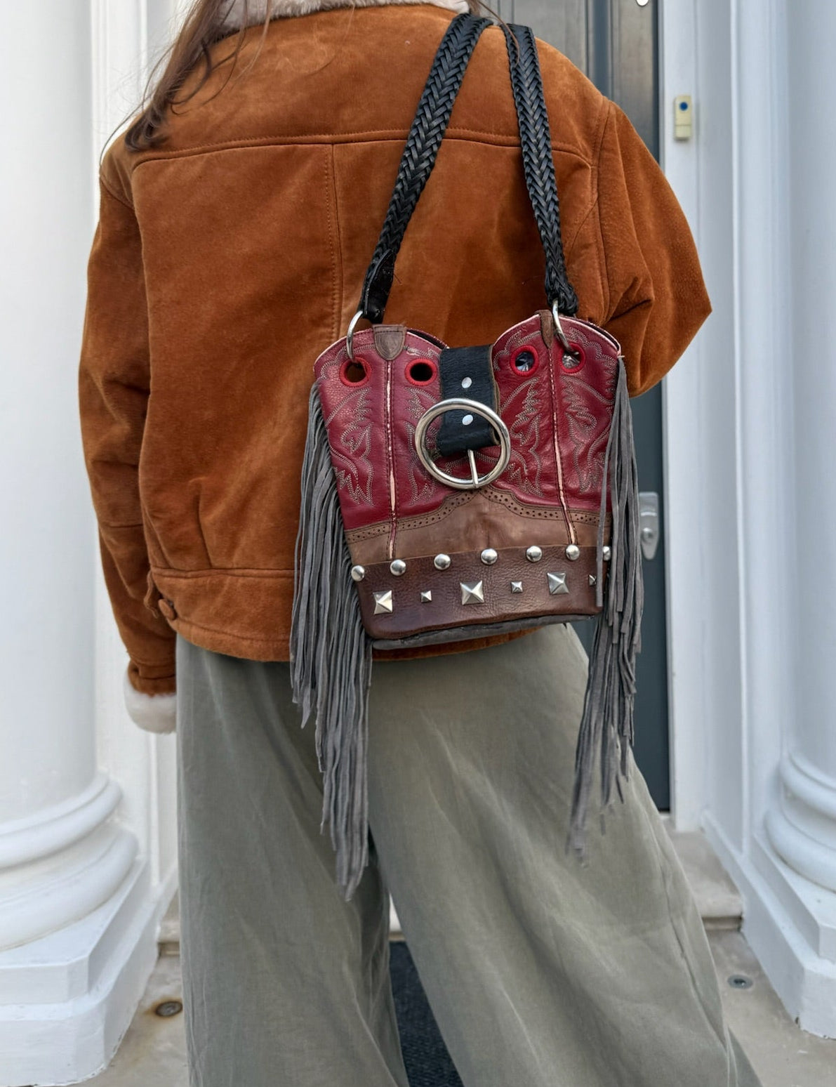 Person wearing a brown jacket and holding a red leather bag with fringe and studs in front of a door.
