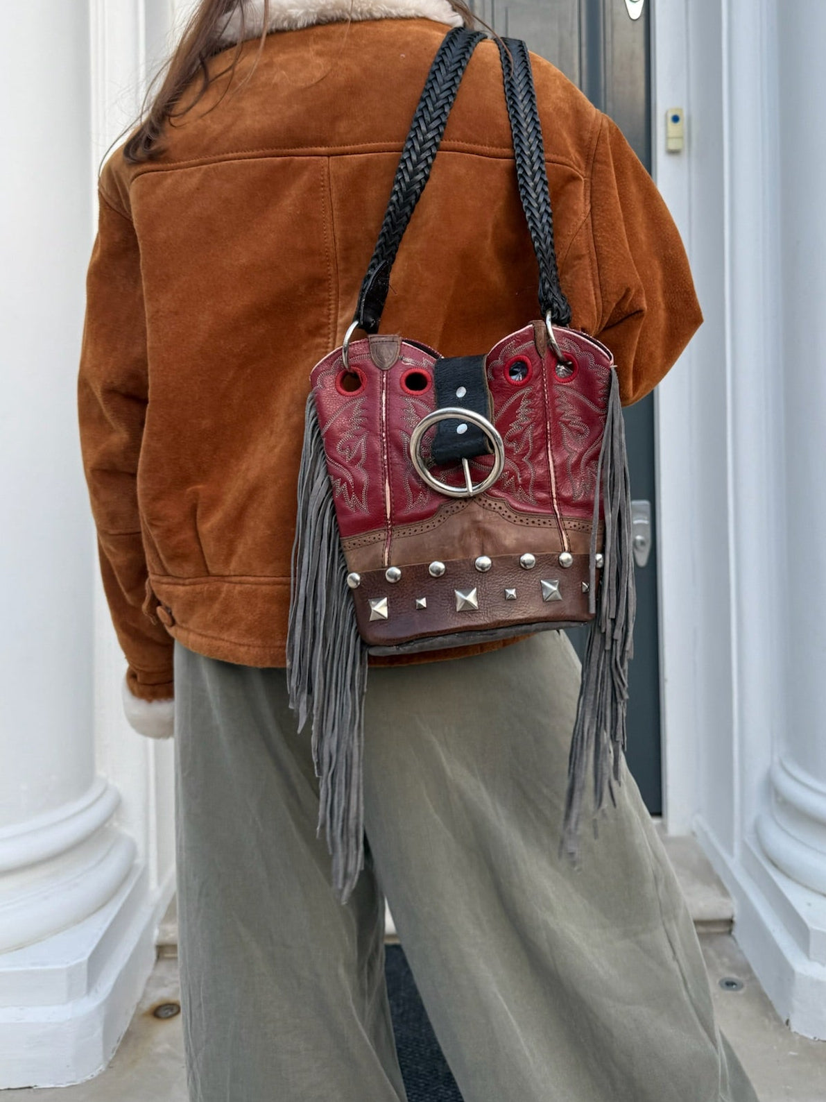 Person wearing a brown jacket and holding a red leather bag with fringe and studs in front of a door.