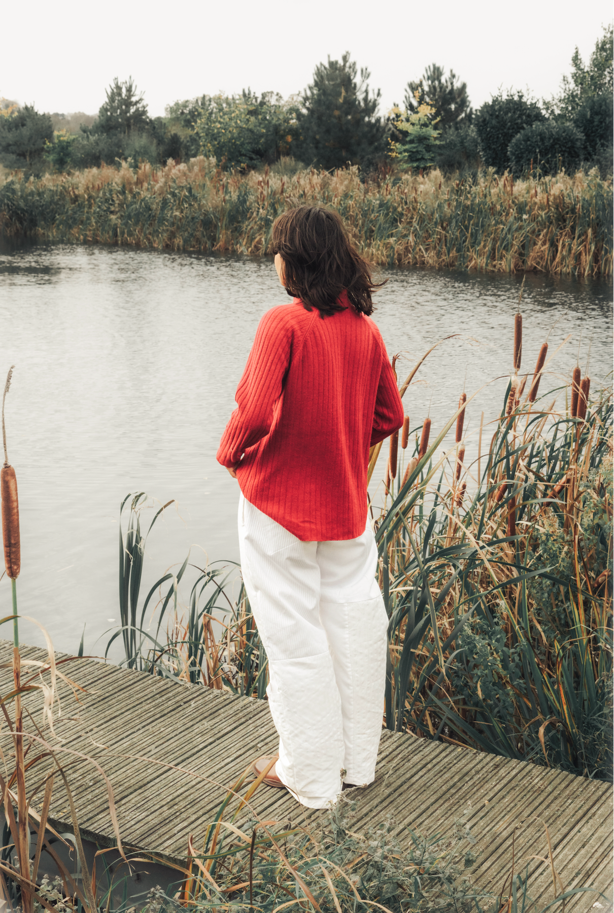 Person standing on a wooden dock by a lake with reeds and trees in the background